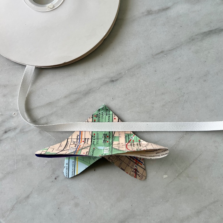 A spool of white ribbon and a folded paper star made from a colorful map rest on a light marble surface, offering inspiration for what to do with old maps. The ribbon stretches across the scene and threads through the star.
