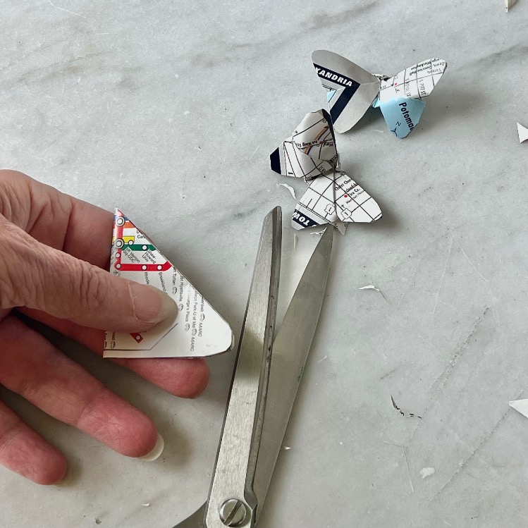 A hand holds a piece of paper with a map design—perfect inspiration for what to do with old maps—next to silver scissors and butterfly shapes crafted from similar map paper on a white surface.