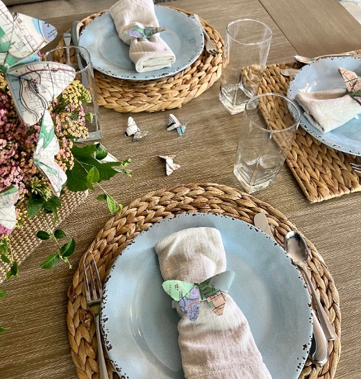 A rustic table setting with woven placemats, pale blue plates, linen napkins decorated with origami butterflies—perfect for those wondering what to do with old maps—and clear glasses. A floral centerpiece and more map butterflies grace the wooden table.