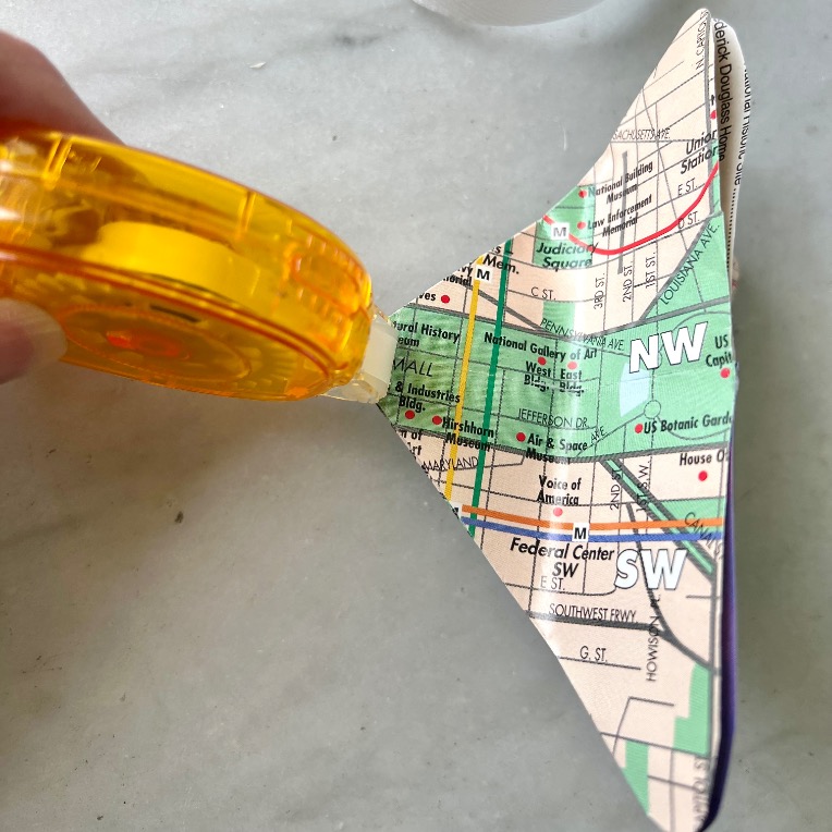 A hand holds a yellow tape dispenser, applying tape to a triangular piece of paper printed with a map featuring streets and landmarks in Washington, D.C.—a creative example of what to do with old maps.