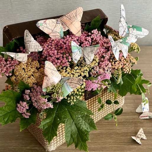 A woven basket filled with pink and yellow flowers and green leaves, decorated with origami butterflies made from old maps—showing a creative idea for what to do with old maps—as more butterflies perch nearby on a wooden surface.