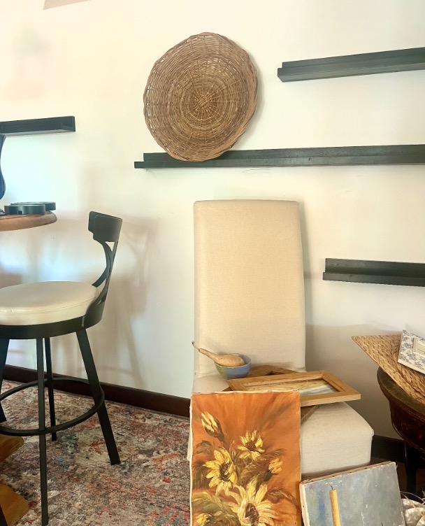 A beige chair with a tray and wooden spoon sits by a wall featuring picture ledge styling with empty black shelves and a woven basket hanging above. A painting of sunflowers leans against the chair on a patterned rug.