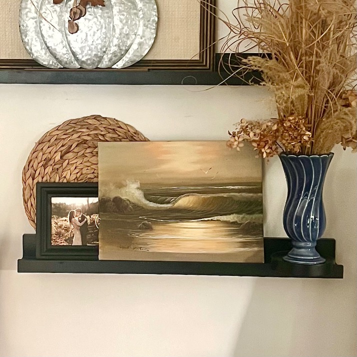 A black shelf showcases picture ledge styling with a framed photo, a round wicker mat, a painting of ocean waves at sunset, and a blue vase with dried flowers against a light-colored wall.