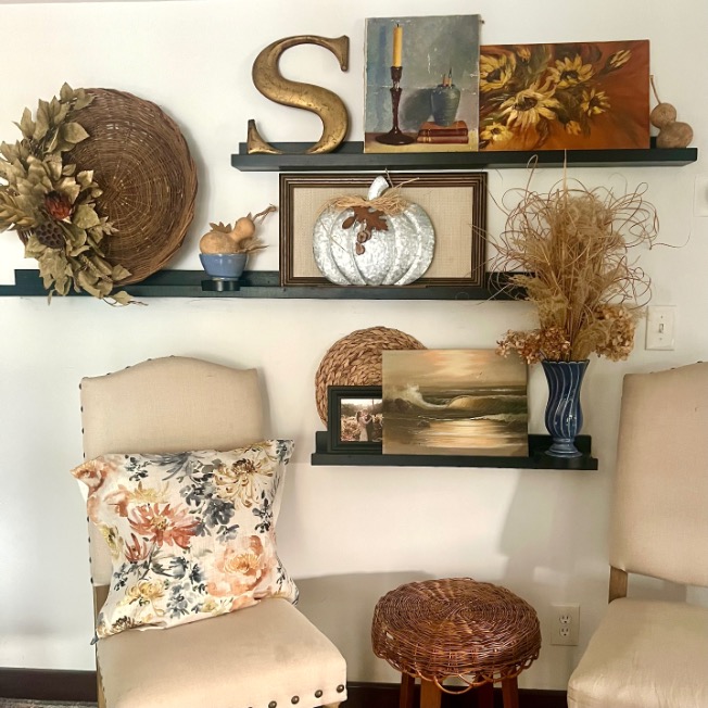 Two beige chairs and a wicker stool sit in front of shelves with fall-themed decor, showcasing picture-ledge-styling with artwork, a white pumpkin, dried plants in a blue vase, a gold "S," basket, and floral pillow.