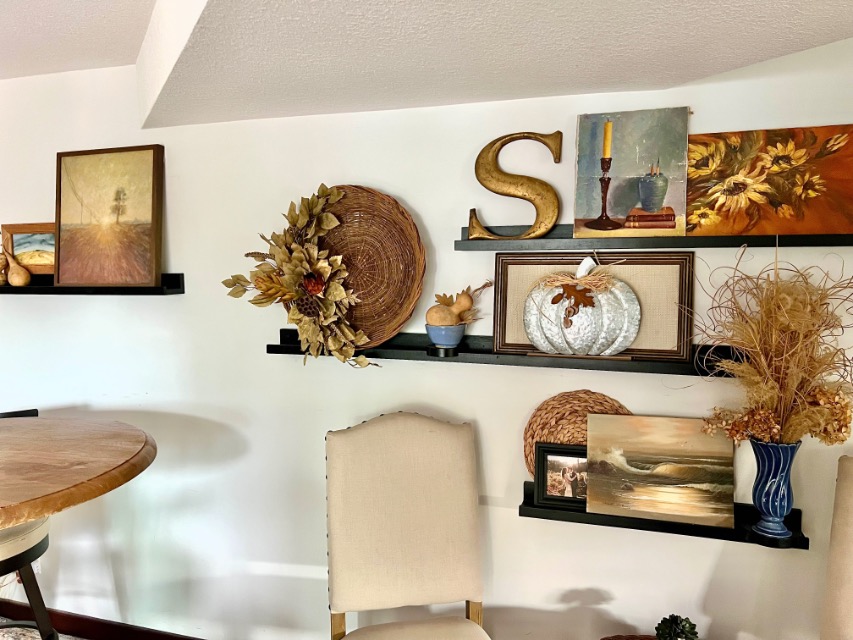Wall shelves showcase picture-ledge-styling with autumn decor—decorative plates, art prints, a gold "S," woven baskets, a pumpkin, dried flowers in a vase, and a beige chair below—creating a cozy and rustic atmosphere.
