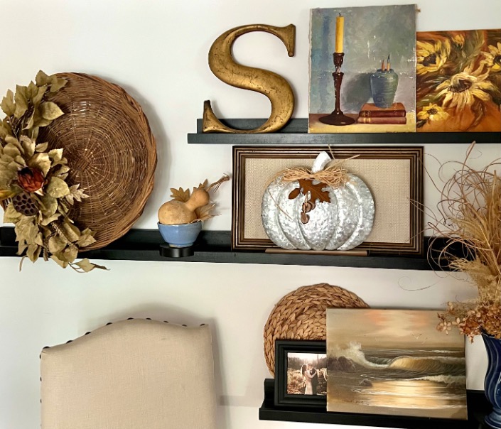 Two black picture ledge shelves display autumn-themed decor, including a gold “S,” a large woven plate with gold accents, a silver pumpkin, framed art, a candle painting, dried flowers, and a round wicker placemat for perfect picture-ledge-styling.