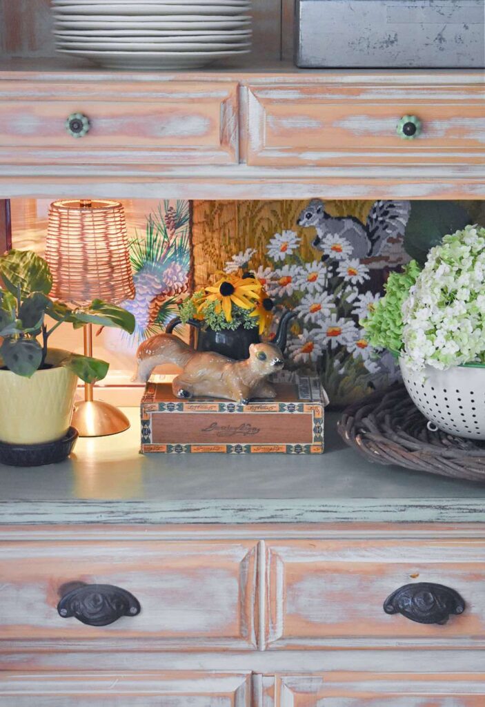 A rustic wooden shelf displays a lamp, potted plants, a ceramic dog figurine on a vintage box, artificial sunflowers, and a white colander with hydrangeas, with a squirrel painting in the background.