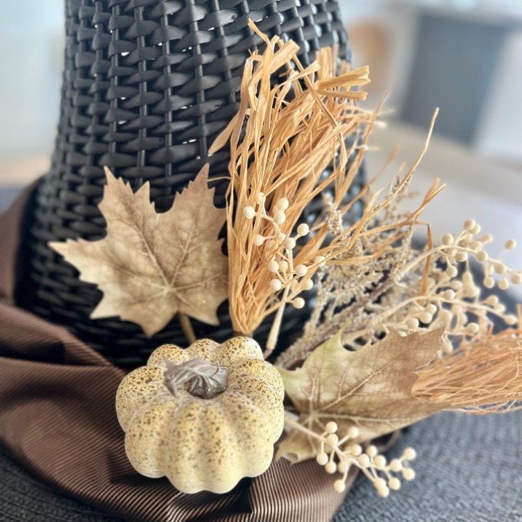 A close-up of a woven black hat decorated with dried beige leaves, wheat, berries, and a small cream-colored pumpkin, evoking an autumn or harvest theme.