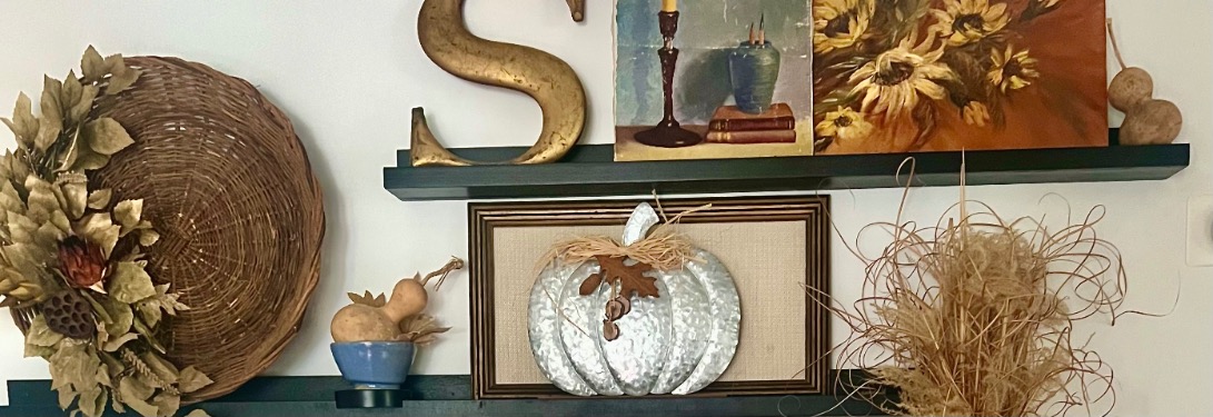 Picture-ledge-styling-hero A decorative picture-ledge-styling arrangement features a woven wreath with gold leaves, a large gold letter "S," a sunflower painting with candles, a metal pumpkin, gourds in a blue bowl, and straw accents on black shelves.