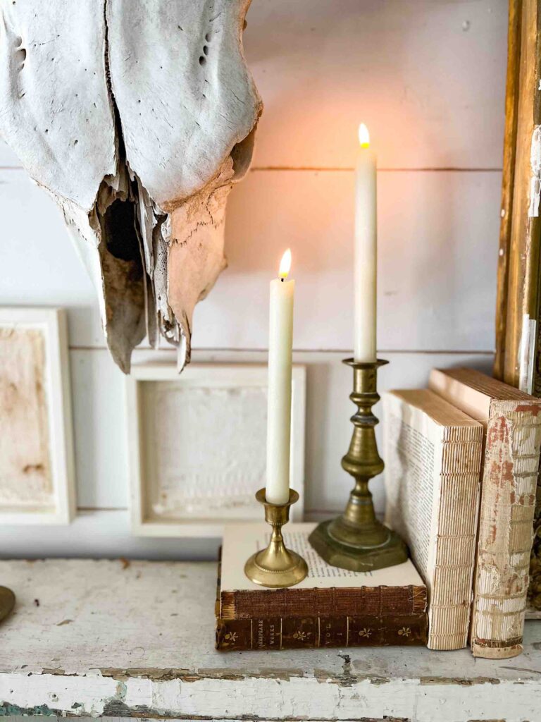 Two lit white candles in brass holders stand on vintage books next to a large animal skull. The scene is set against a white wall with framed art and a rustic, weathered surface.