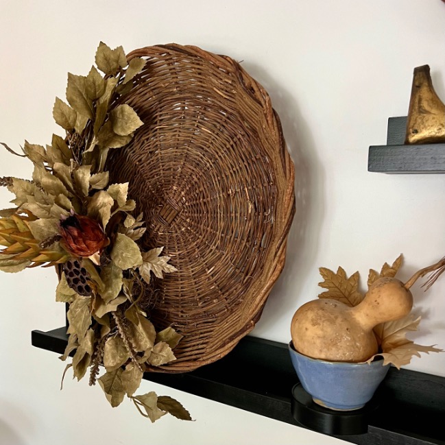 A round wicker basket with dried leaves and flowers hangs above a black picture ledge, styled with a blue bowl holding dried gourds and leaves. Another shelf displays a small brass pear, adding charm to the picture-ledge-styling.