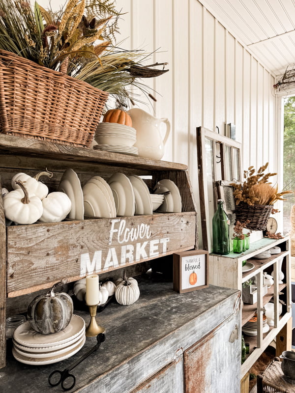 A rustic kitchen display with a wooden hutch labeled "Flower Market," white dishes, white and gray pumpkins, candles, and a wicker basket of dried plants. A shelf with more decor and baskets is in the background.