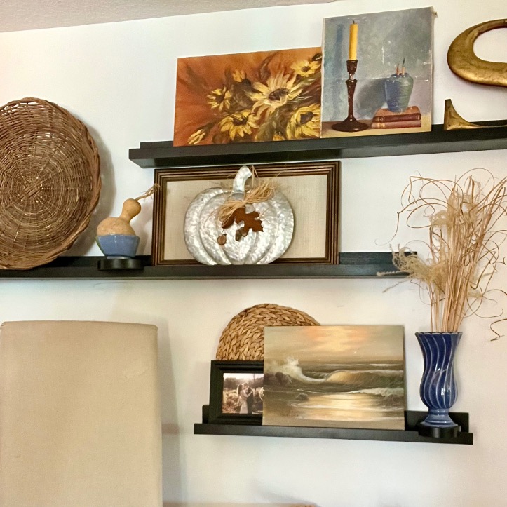 Three wall shelves showcase picture-ledge-styling with woven baskets, framed art of sunflowers and the ocean, a metal pumpkin, a blue vase with dried grass, a yellow candle, and decorative objects against a light-colored wall.