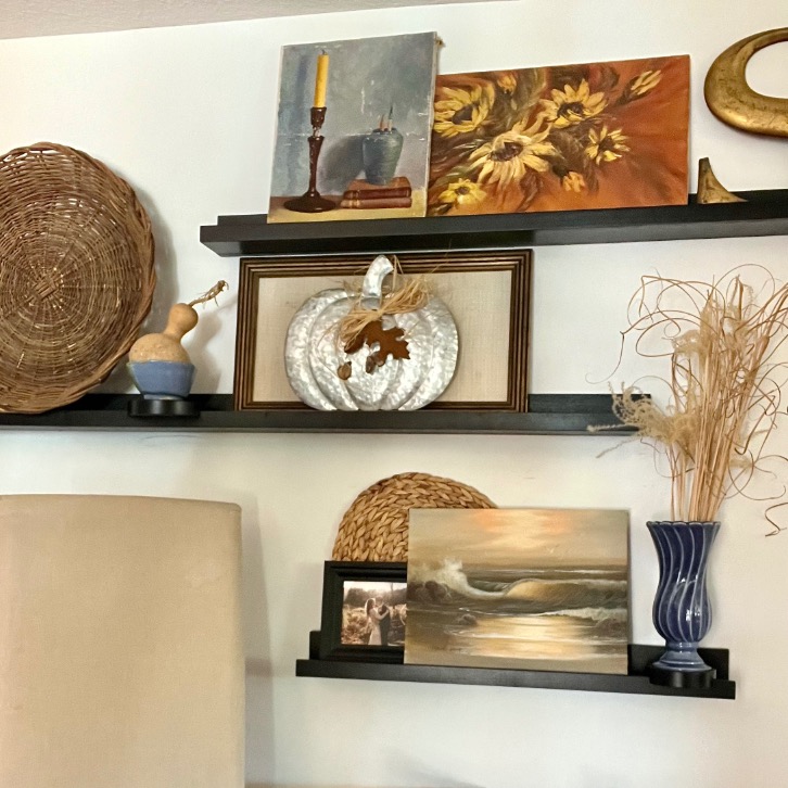 Three black wall shelves showcase picture-ledge-styling with woven baskets, framed art of a sunflower and ocean scene, a silver pumpkin ornament, a gourd, and dried grasses in a blue vase against a white wall.
