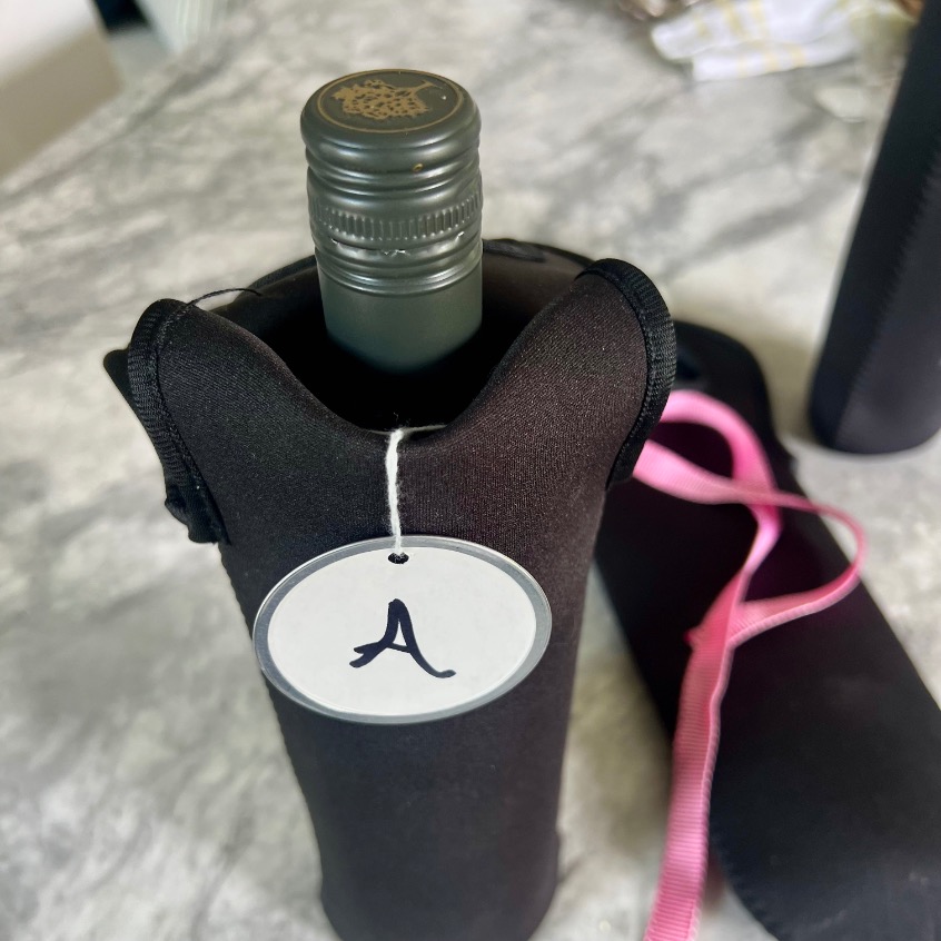 A wine bottle with a metal screw cap is wrapped in a black fabric cover, perfect for a blind wine tasting party. A round white tag with the letter "A" is attached, while a pink ribbon and another similarly covered bottle sit on the gray surface.