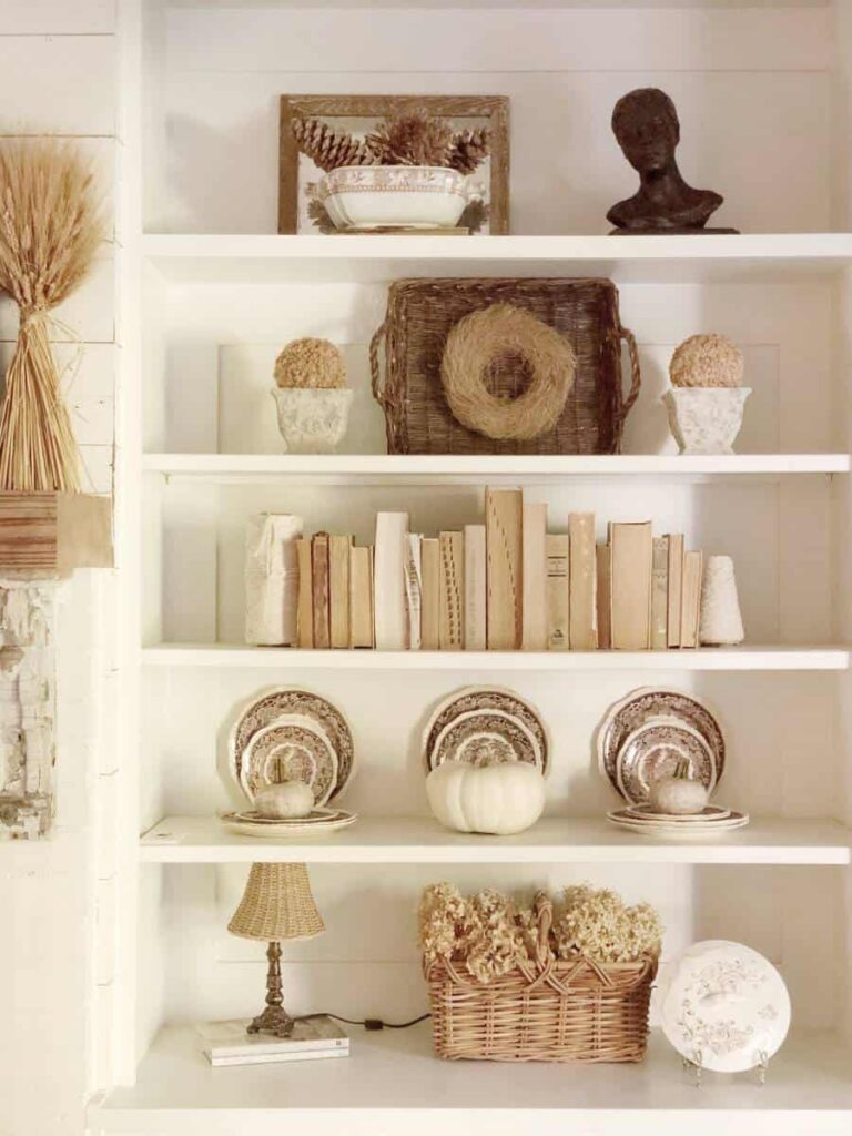 A white shelving unit showcases picture ledge styling with neutral-toned decor: woven basket, books, bust sculpture, plates, dried flowers, a small lamp, and a white pumpkin arranged in rustic farmhouse style.