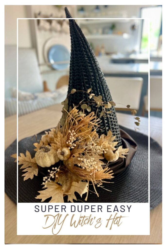 A black witch’s hat, perfect for DIY witch hat decor, is adorned with dried leaves, small pumpkins, and flowers on a table. Text reads: “Super Duper Easy DIY Witch’s Hat.” A cozy, modern living room is in the background.