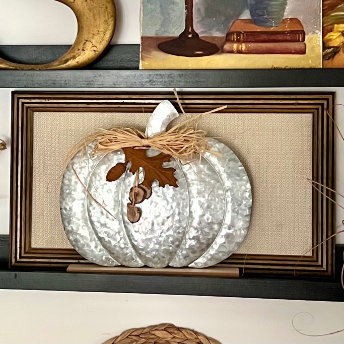 A decorative silver metal pumpkin with a raffia bow and brown leaves sits on a shelf, set against a textured background and surrounded by framed artwork—perfect inspiration for picture ledge styling with autumn decor accents.