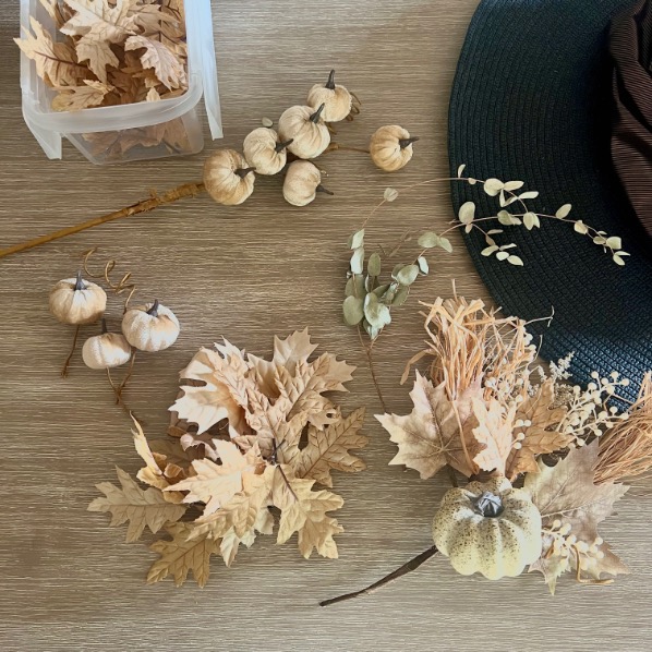 A tabletop displays artificial beige pumpkins, dried leaves, and foliage, with a plastic container holding more pumpkins, and part of a dark green hat visible in the corner.