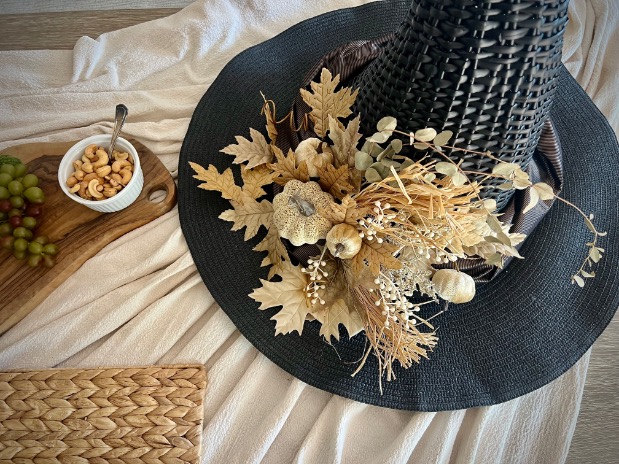 A black wicker witch hat decorated with dried leaves and flowers sits on a cream cloth beside a wooden board with grapes and a small bowl of cashews. A woven mat is partially visible in the corner.