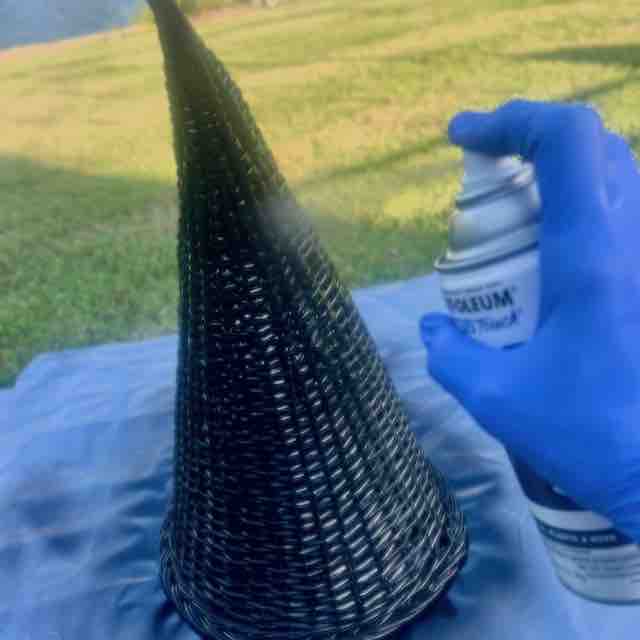 A person wearing a blue glove sprays black paint onto a cone-shaped wicker object outdoors, with grass in the background.