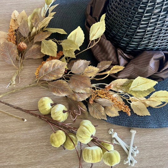 A black woven hat with a brown ribbon sits on a wooden surface next to artificial autumn leaves, beige flowers, green pod decorations, and small white plastic bones.