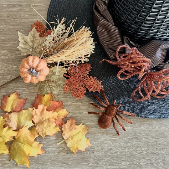 A decorative black hat, artificial autumn leaves, a faux pumpkin, a small broom, a brown ribbon, and a fake spider arranged on a wooden surface, evoking a fall or Halloween theme.