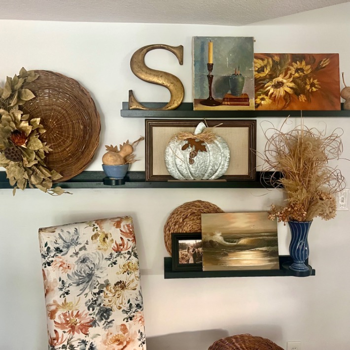 Wall shelves showcase picture-ledge-styling with a gold letter “S,” sunflower paintings, candles, a white pumpkin frame, woven baskets, floral and abstract art, a blue vase with dried grasses, and small decorative birds.