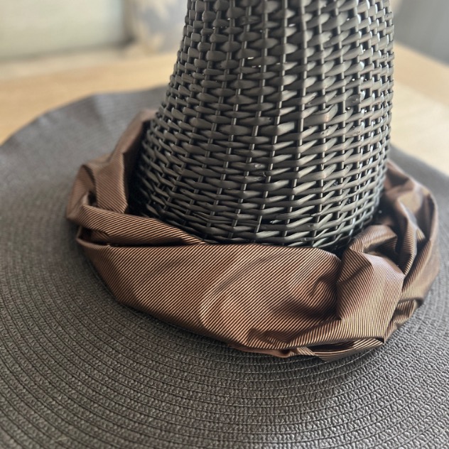 A close-up of a woven, dark brown witch hat with a wide brim and a brown fabric band around the base of the cone, placed on a light-colored surface.