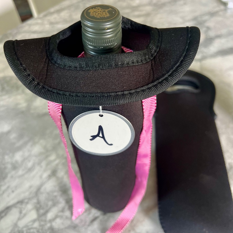 A bottle in a black neoprene wine tote with a pink ribbon and a round tag featuring the letter "A," perfect for a blind wine tasting party, sits on a marble surface next to an empty matching bottle holder.