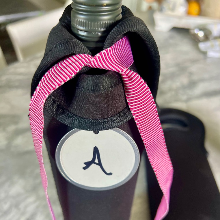 A bottle covered in a black sleeve with a pink-striped ribbon at the neck and a round tag featuring the letter "A" sits on a marble countertop—perfect for a blind wine tasting party.