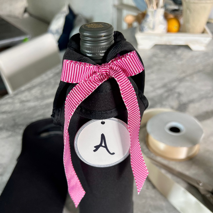 A bottle wrapped in black fabric, perfect for a blind wine tasting party, with a pink and white striped ribbon tied in a bow around the neck. A round white tag with the letter "A" hangs from the ribbon. In the background are a spool of ribbon and other items.