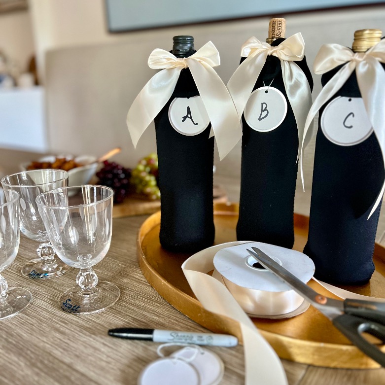 Three wine bottles wrapped in black fabric and labeled A, B, and C with ivory ribbon bows are displayed on a tray with empty wine glasses, a marker, white ribbon, tags, and scissors—perfect for a blind wine tasting party.