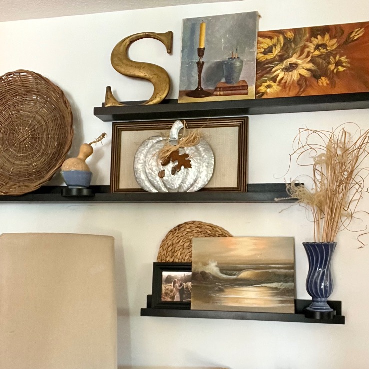 Three black wall shelves showcase picture-ledge-styling, featuring a gold letter "S," baskets, a sunflower painting, a pumpkin ornament, a blue vase with dried grass, and ocean artwork—all artfully arranged above a beige lampshade.