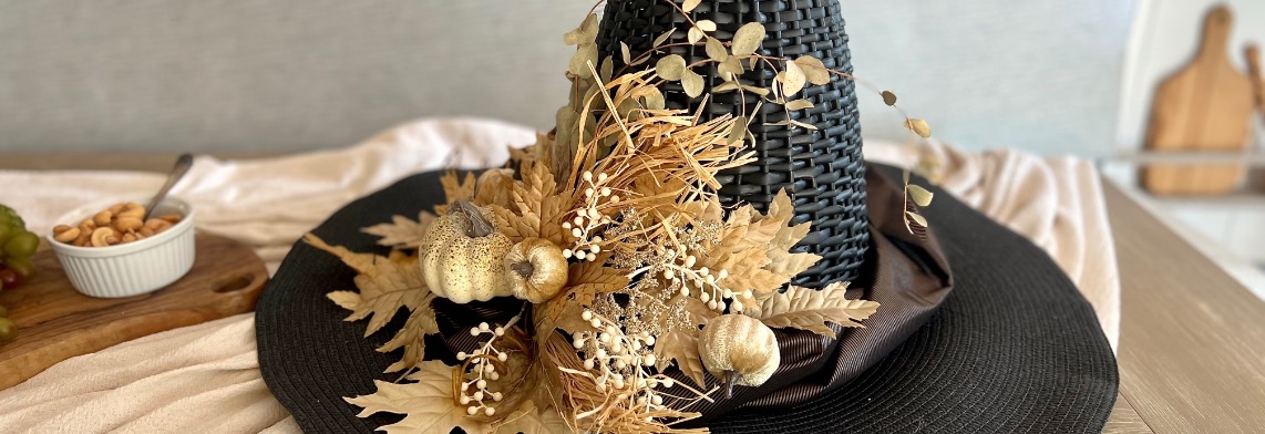 witch-hat-hero A black woven hat decorated with dried leaves, small pumpkins, and other autumn-themed ornaments sits on a table next to a wooden board with nuts and grapes.