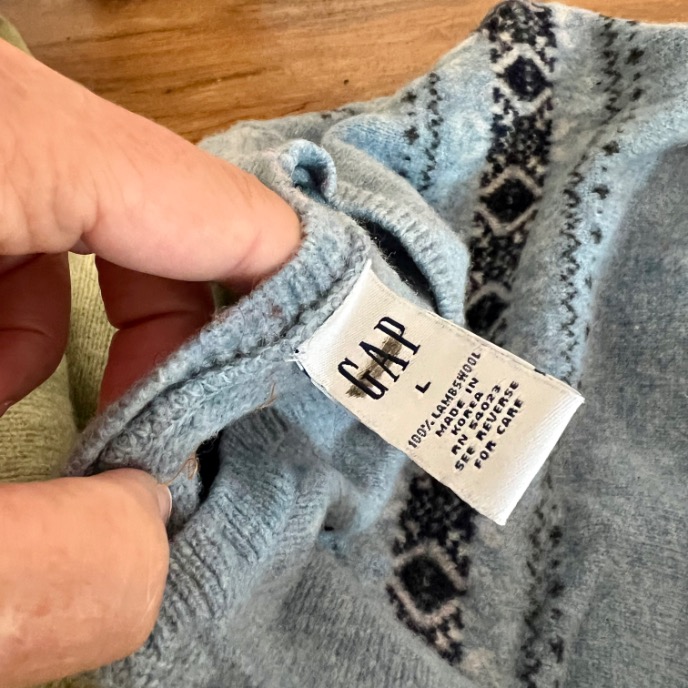 A hand holds a light blue sweater with a white GAP label showing size L and washing instructions. The sweater has a subtle dark blue patterned design near the collar, perfect for anyone looking to upcycle an old sweater into something new.