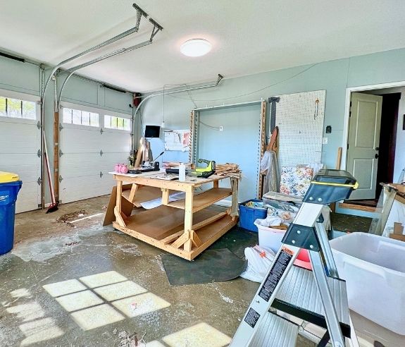 A sunlit garage with an open door, a large wooden workbench, a stepladder, storage bins, a pegboard with tools, and miscellaneous items scattered across the floor.