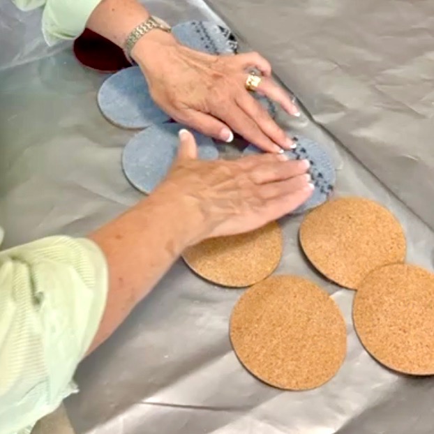 Two hands smooth out round denim fabric pieces, possibly upcycled from an old sweater, beside several cork rounds on a table covered with shiny cloth. The person wears a light green shirt and a watch.