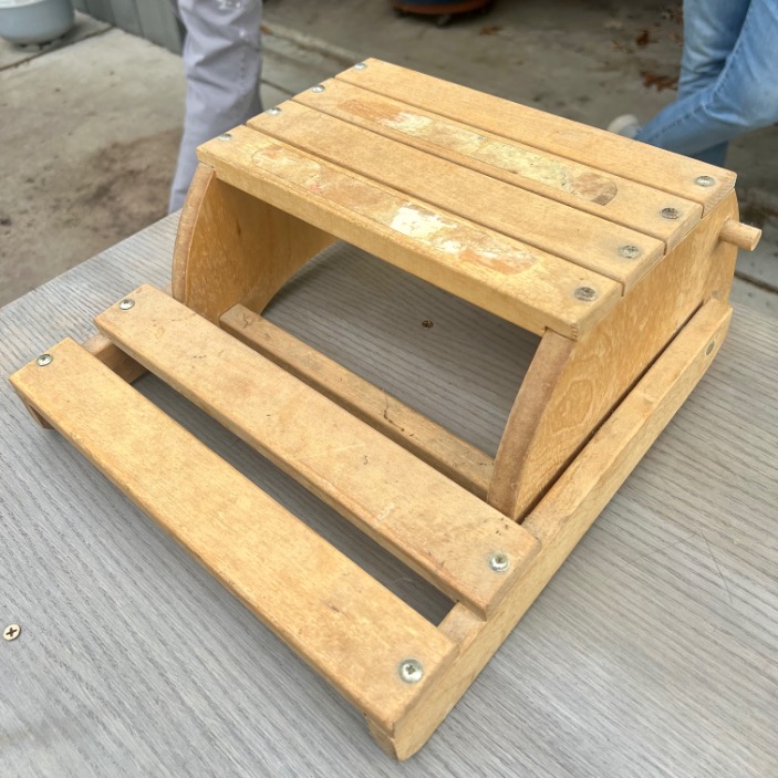 A small, unfinished wooden structure with a hinged, slatted top and sides, sitting on a gray table. A person's legs are visible in the background. The structure appears to be a handcrafted project.