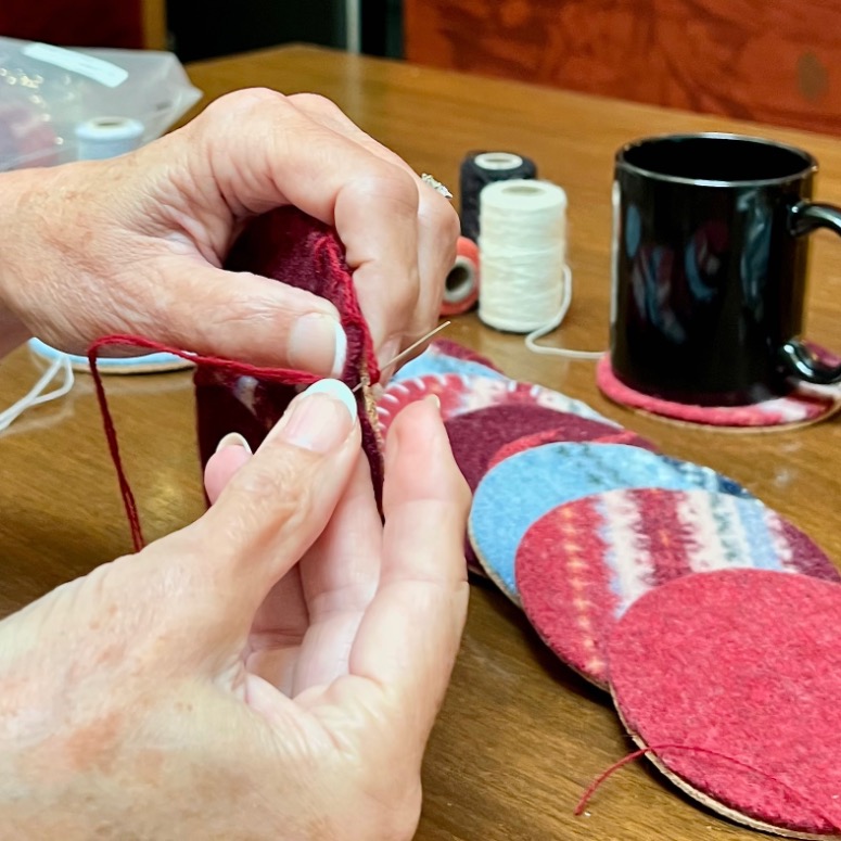 Close-up of hands sewing a red felt circle with a needle and yarn—an easy way to upcycle an old sweater. Spools of thread, more felt circles in red and blue, and a black mug on a coaster sit on the wooden table.