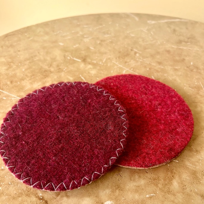 Two round felt coasters, upcycled from an old sweater—one burgundy with visible white zigzag stitching around the edge and one red with no visible stitching—are placed on a beige marble surface.