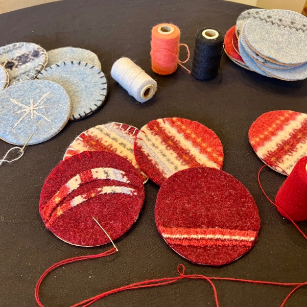 Several round pieces of colorful fabric, mostly red and patterned, are being hand-stitched on a table—an ideal way to upcycle old sweater scraps. Spools of thread and completed fabric circles are also visible nearby.