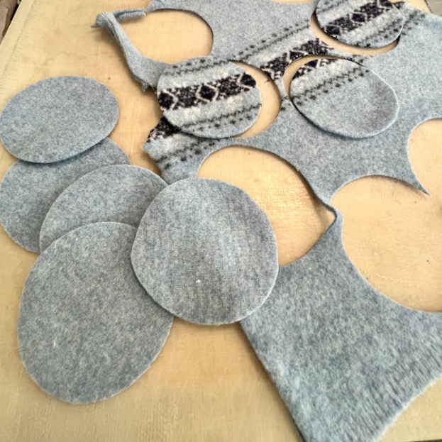 Several light blue felt circles, upcycled from an old sweater, are cut out from patterned fabric lying on a wooden surface. The fabric features dark geometric designs and various circular cutouts.