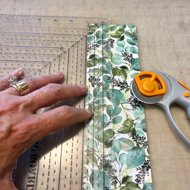 A hand holds a clear ruler over a strip of green leafy fabric, preparing to cut it with a rotary cutter on a beige surface.
