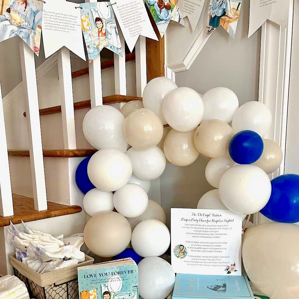 A display by a staircase with white and blue balloons, children’s books, a basket of diapers, and a banner of illustrated flags hangs above—a festive baby shower or children’s party setup, perfect for showing off your latest diy-stool-makeover.