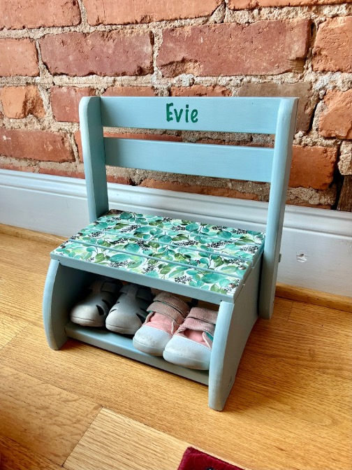 A small light blue wooden chair with "Evie" on the backrest, featuring a floral-patterned seat and a lower shelf for toddler shoes, sits against a brick wall—a charming diy-stool-makeover.