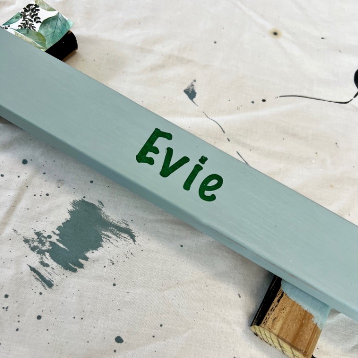 A light blue wooden plank with the name "Evie" painted in green sits on a paint-splattered white surface, supported by two blocks underneath.