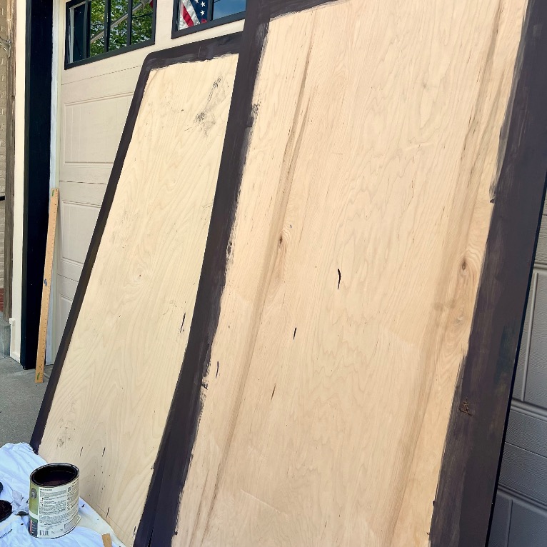 Two large plywood boards, possibly for twin beds to X-long conversions, lean against a garage door, partially painted dark brown along the edges. A paint can and brush rest on a tarp nearby. The garage and part of a brick wall are visible.