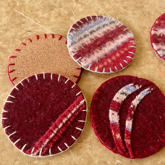 Several handmade, round felt coasters—crafted by upcycling an old sweater—feature shades of red and tan, decorative stitching, and layered patterns, all displayed on a beige surface.