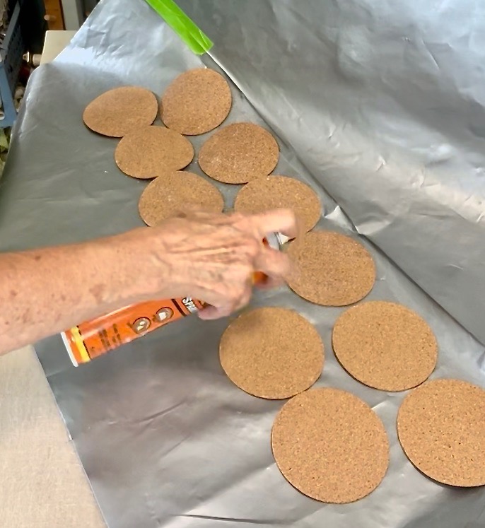 A hand is spraying adhesive onto a row of round cork coasters placed on a sheet of aluminum foil, perfect for an upcycle old sweater project.